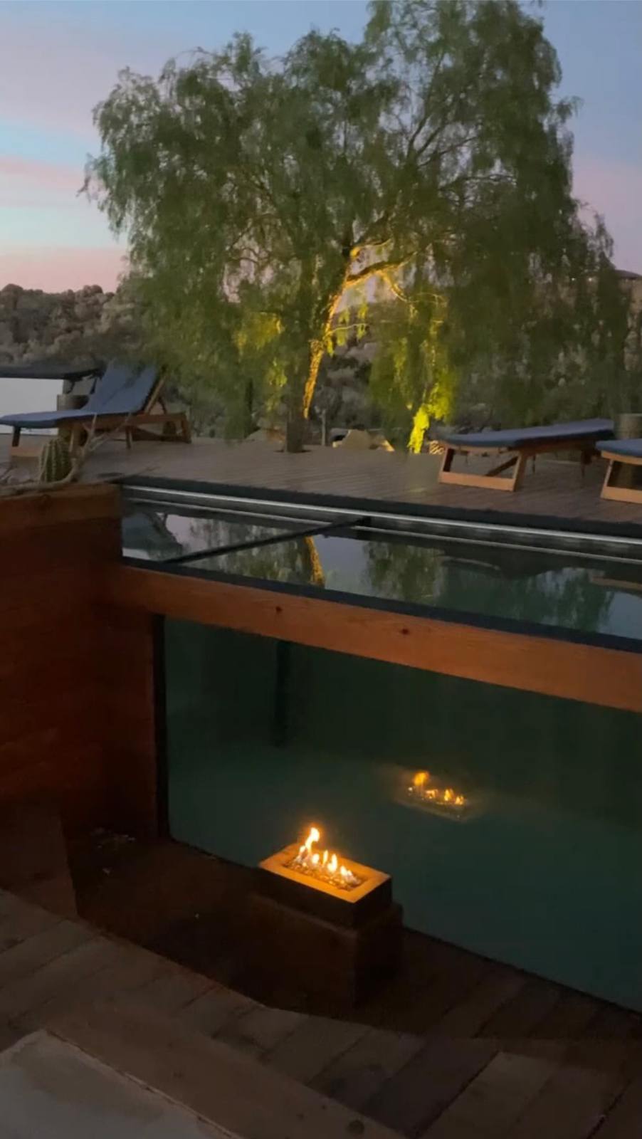 Glass-walled swimming pool with fire feature at dusk on the redwood deck